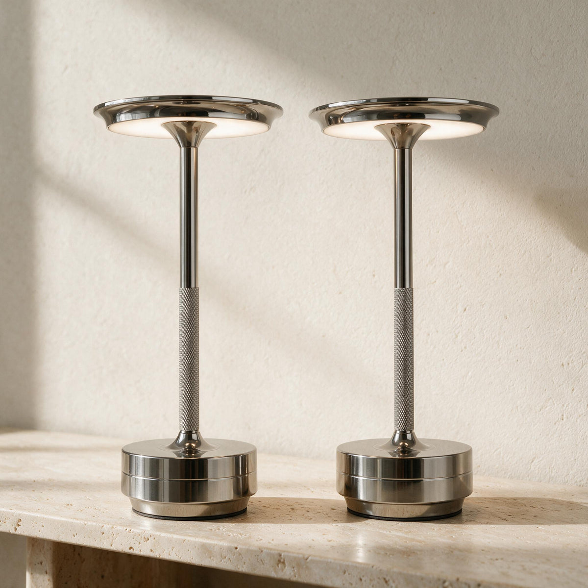 Two silver Cordless Metal Table Lamps casting warm light on a travertine console.